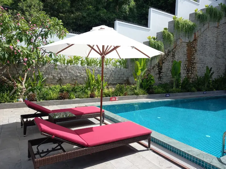 Ghế hồ bơi nhựa giả mây cao cấp ngoài trời tại resort với thiết kế giường tắm nắng, đệm đỏ, dù che nắng sang trọng